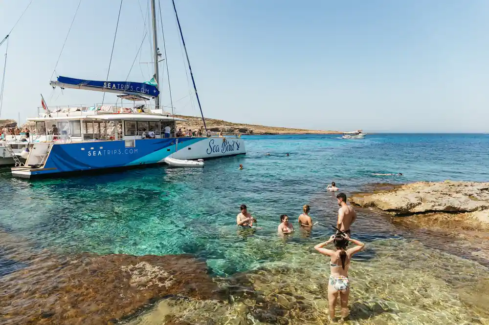 The blue-hulled catamaran anchored off a rocky shore with swimmers in the shallows
