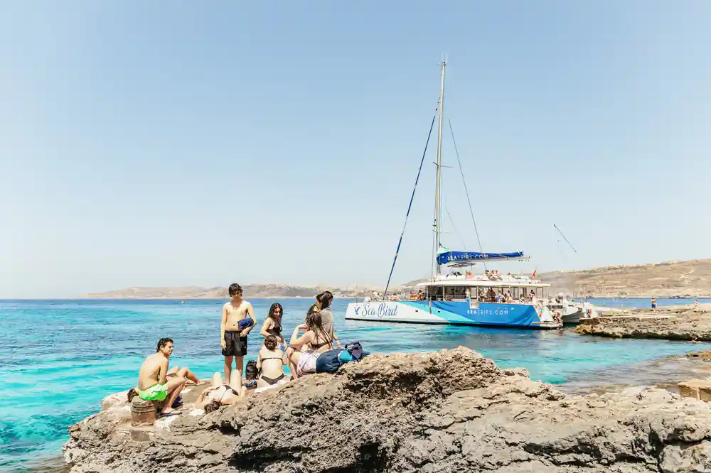 The catamaran anchored off a rocky shore with people gathered on the rock