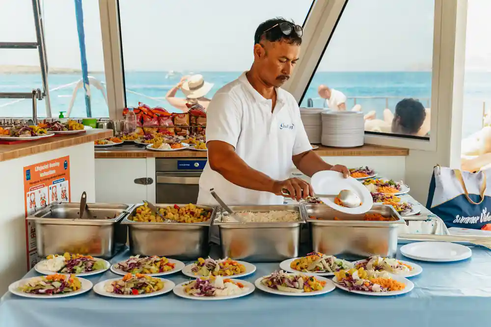 A crew member plating up food from trays at the boat's galley counter